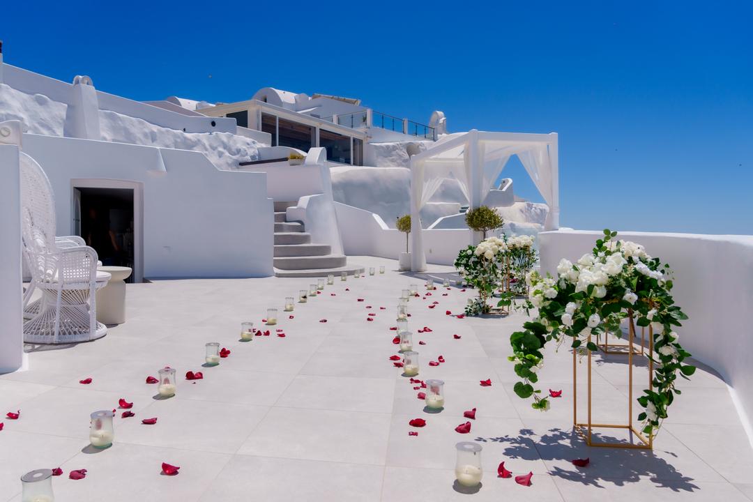 Rose petals and candle glasses laid in a path across an empty white terrace toward a tall stand of white flowers