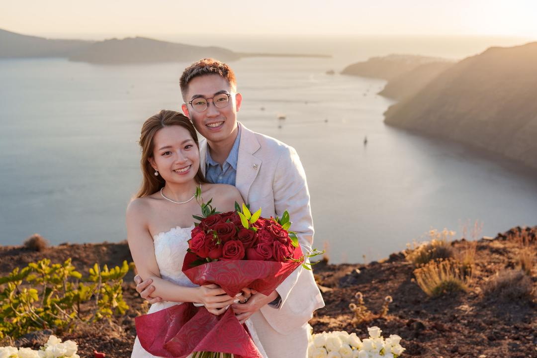 A couple holding a wrapped bouquet of red roses on the rim in warm evening light, white flowers at their feet