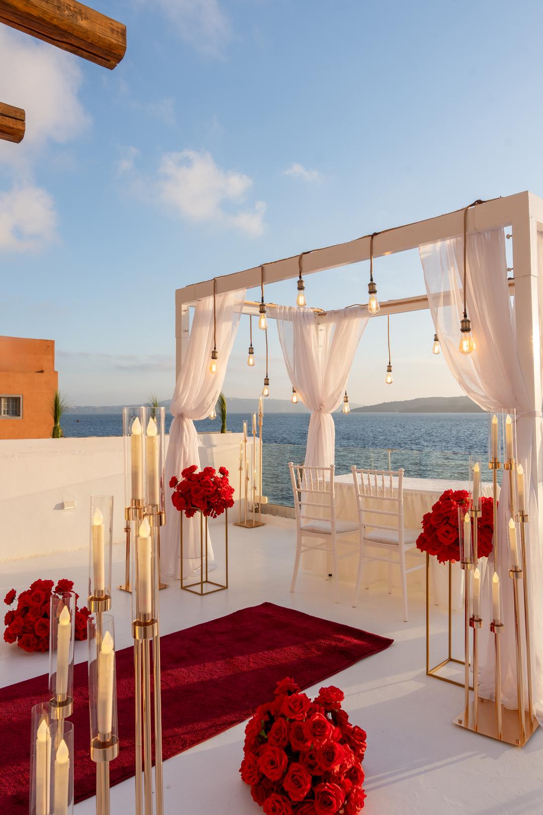 Two pale chairs under a draped canopy with banks of red roses and gold candlesticks, the caldera behind