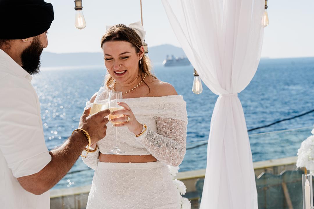 A woman laughing with a glass of champagne under hanging filament bulbs and white drapes, the sea behind