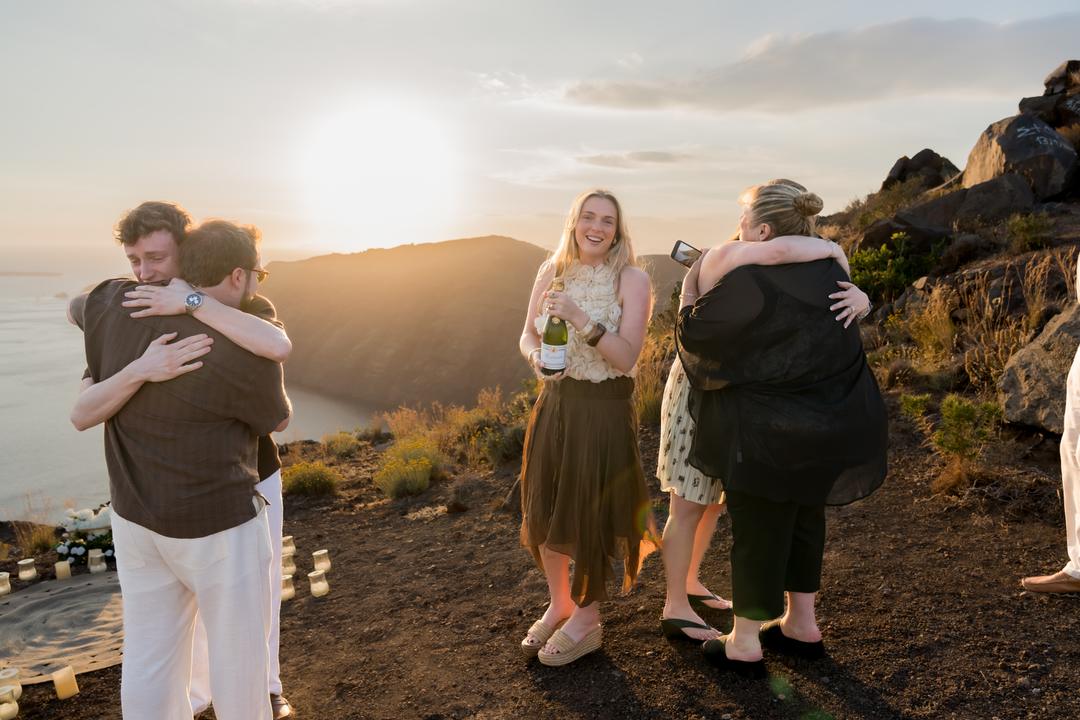 Two pairs of people embracing on a ridge at sunset beside a woman holding a bottle of champagne, a ring of candles behind them