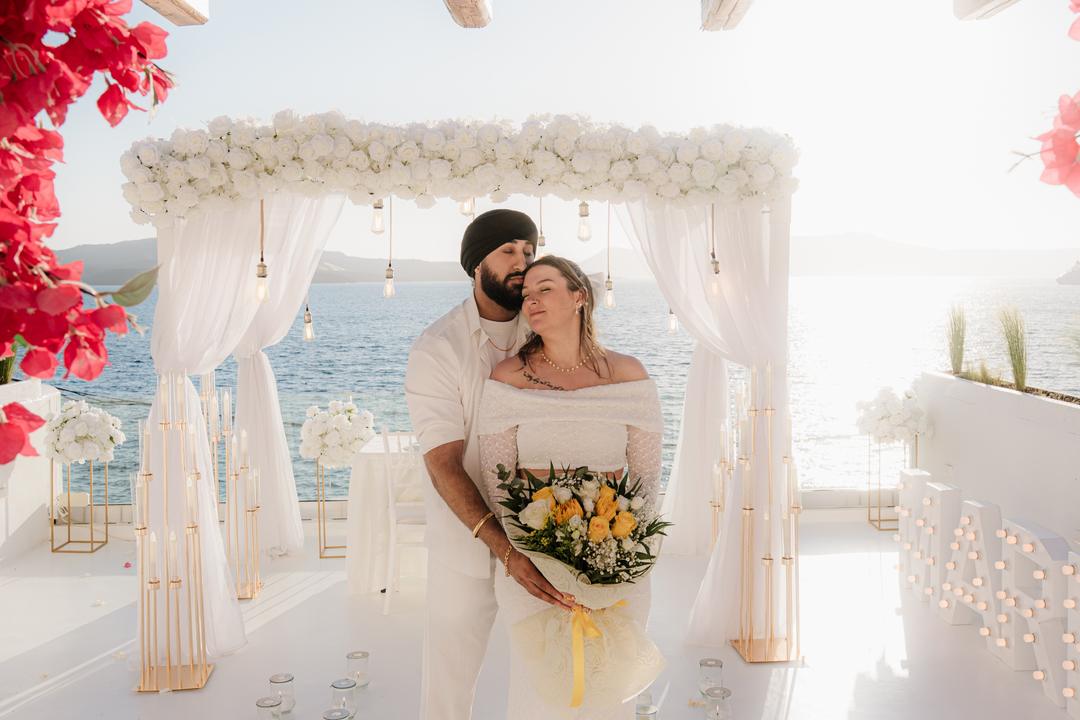 A couple standing under a white floral arch holding a yellow and white bouquet, bougainvillea overhanging the frame and the Aegean behind them