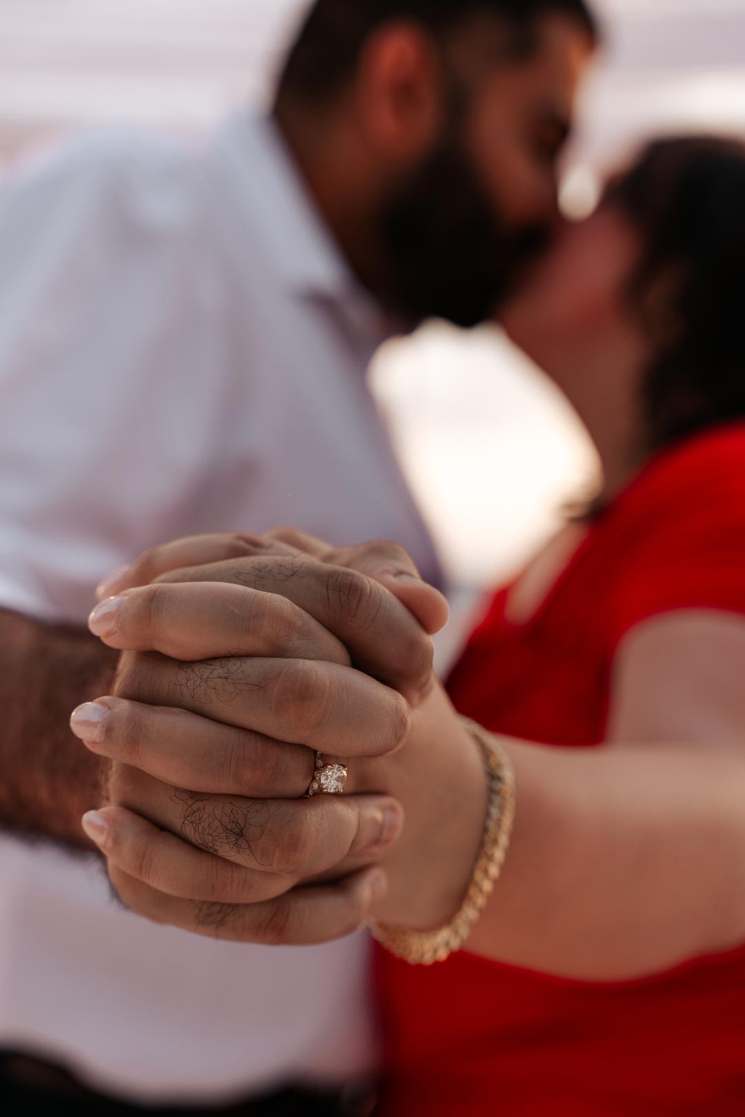 Two pairs of hands clasped together with a new ring on one of them, a kiss out of focus behind