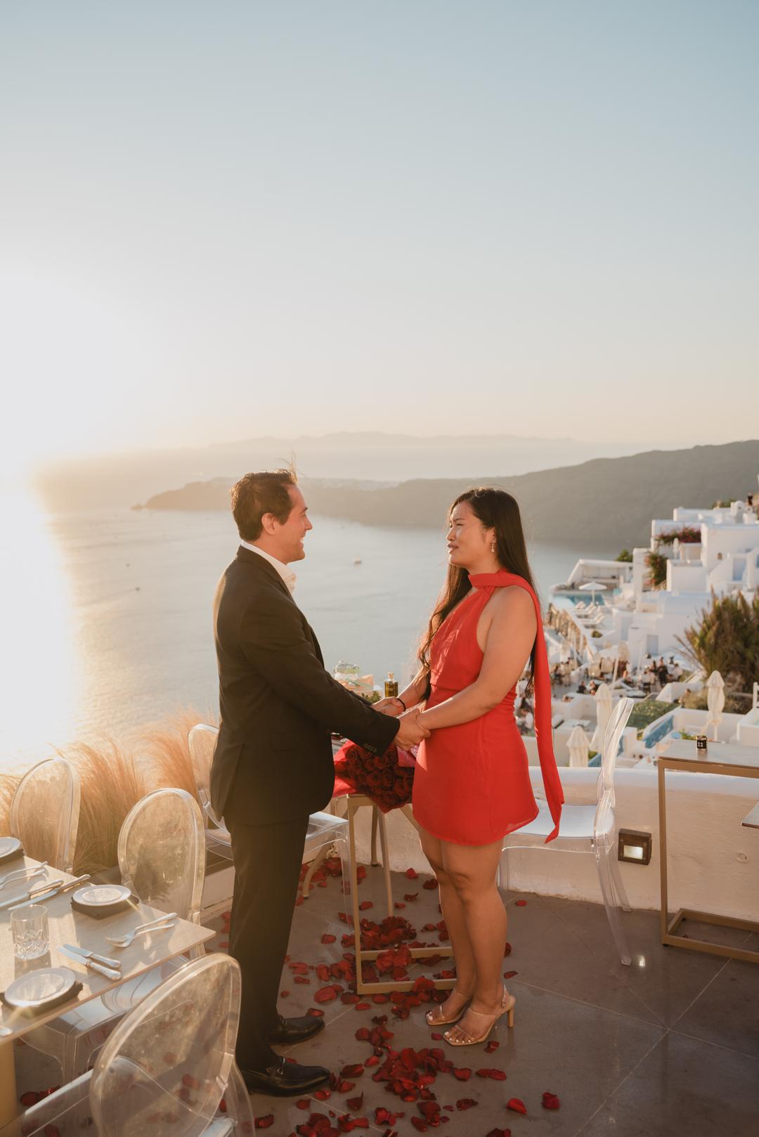 A couple holding both hands facing each other on a terrace strewn with rose petals, the village and the caldera in golden light