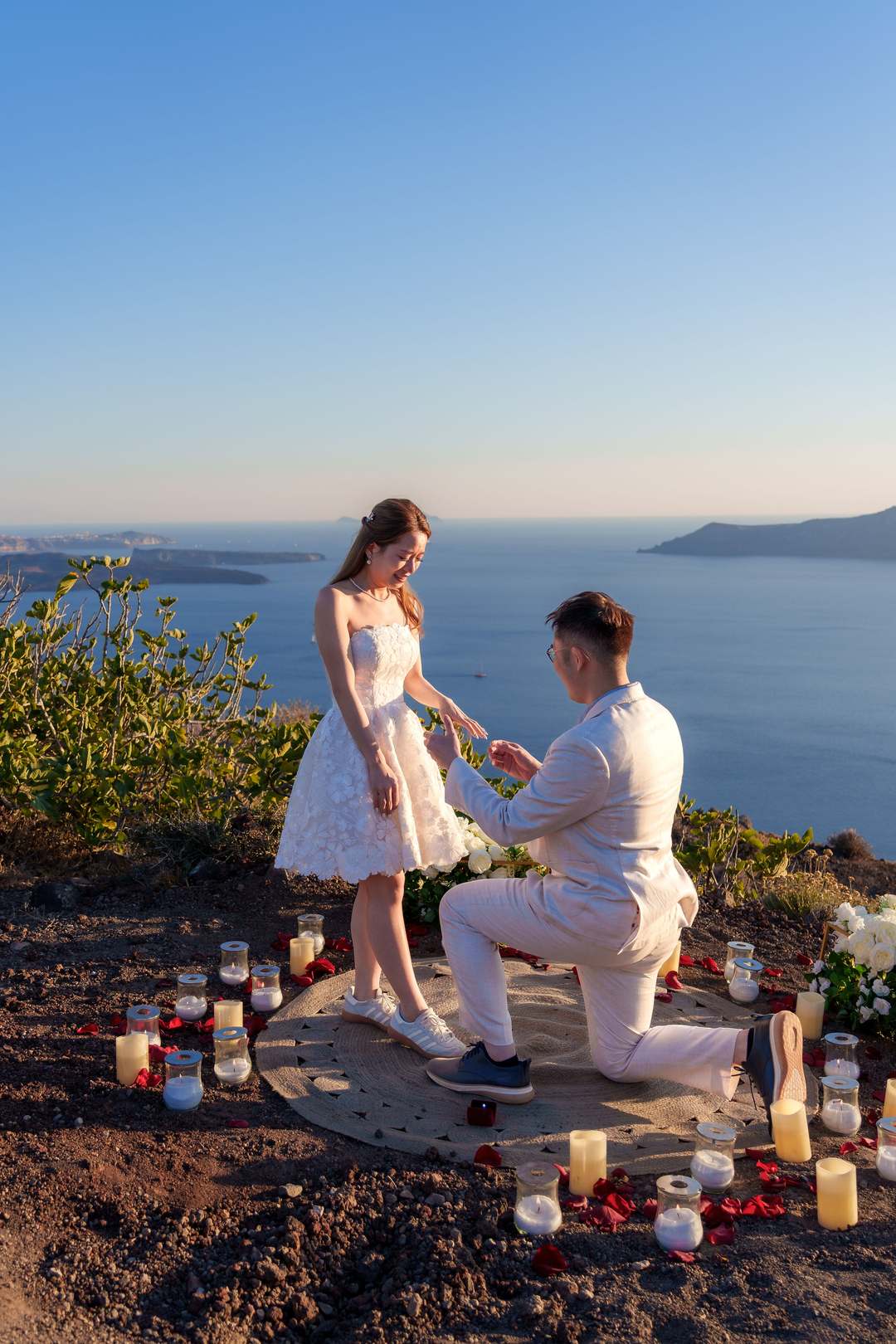 A man on one knee inside a setting of candles and petals on the rim, holding her hand, the sea and the far islands behind