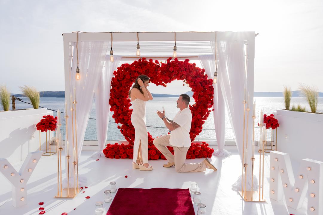 A man on one knee inside a red rose heart arch, a woman with both hands to her face, red carpet underfoot and the sea behind