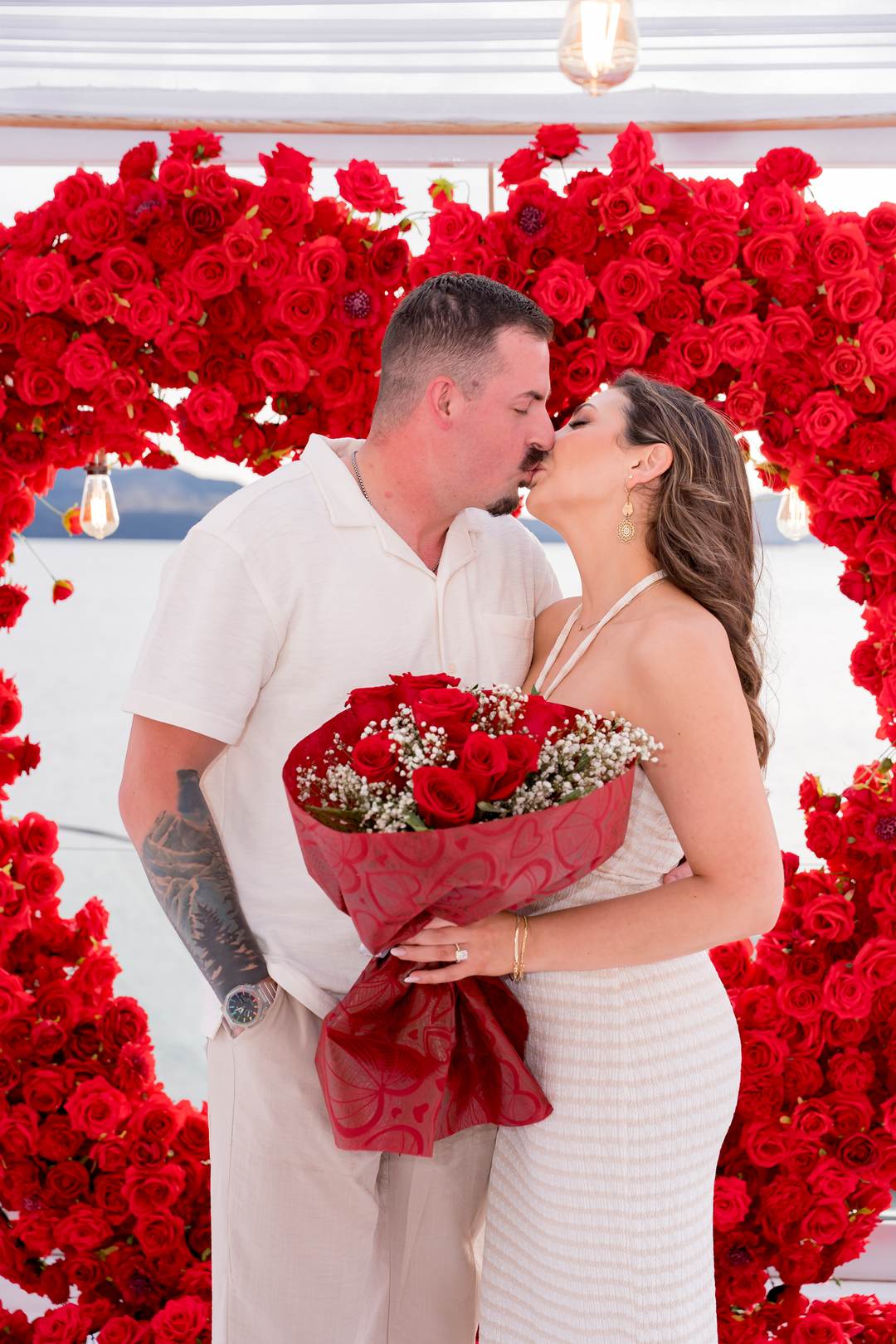 A couple kissing in front of a wall of red roses, holding a bouquet wrapped in red paper