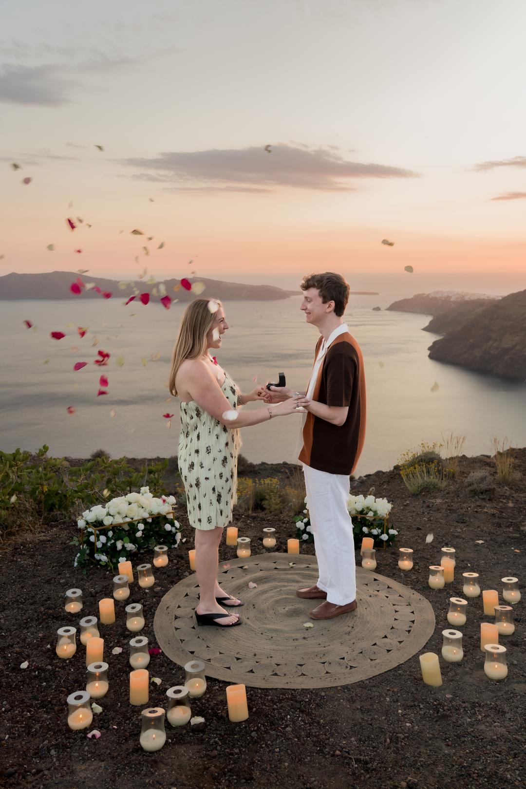 Rose petals falling through the air as a couple stand facing each other on a round rug ringed with candles, a ring box in his hand and the caldera behind