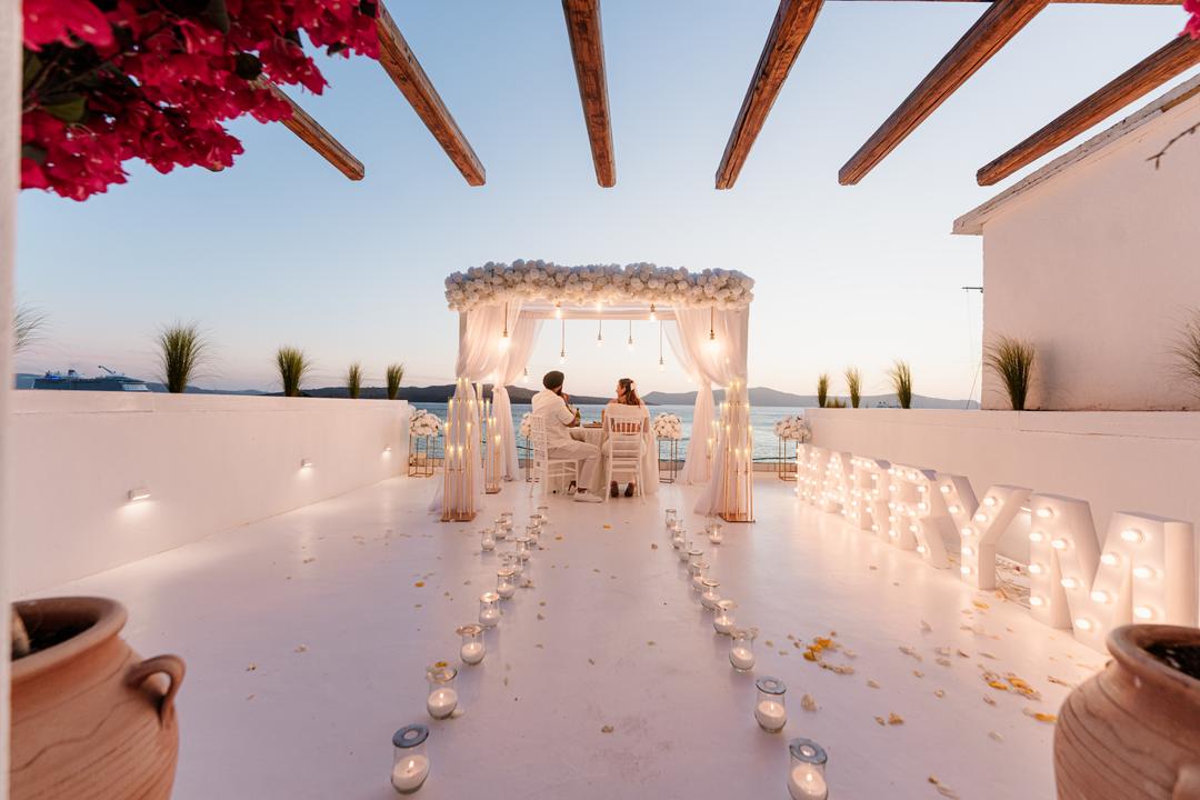 A candle-lined aisle of rose petals crossing a white terrace to a draped arch, lit MARRY ME letters at one side and bougainvillea overhead