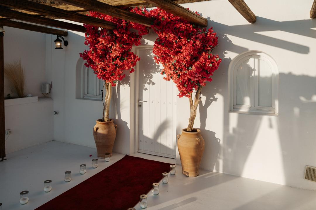 A white venue entrance with two red bougainvillea in terracotta pots and a red carpet lined with candles running to the door