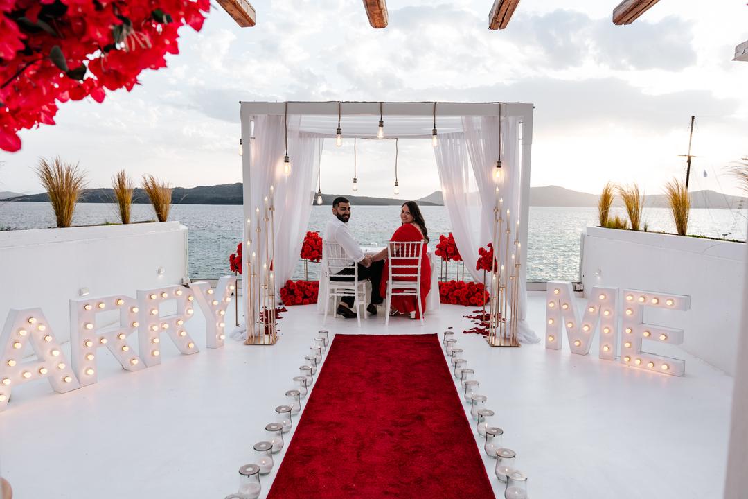 A red carpet running between lit MARRY ME letters to a white draped canopy hung with red roses, a couple seated beneath it and the sea beyond