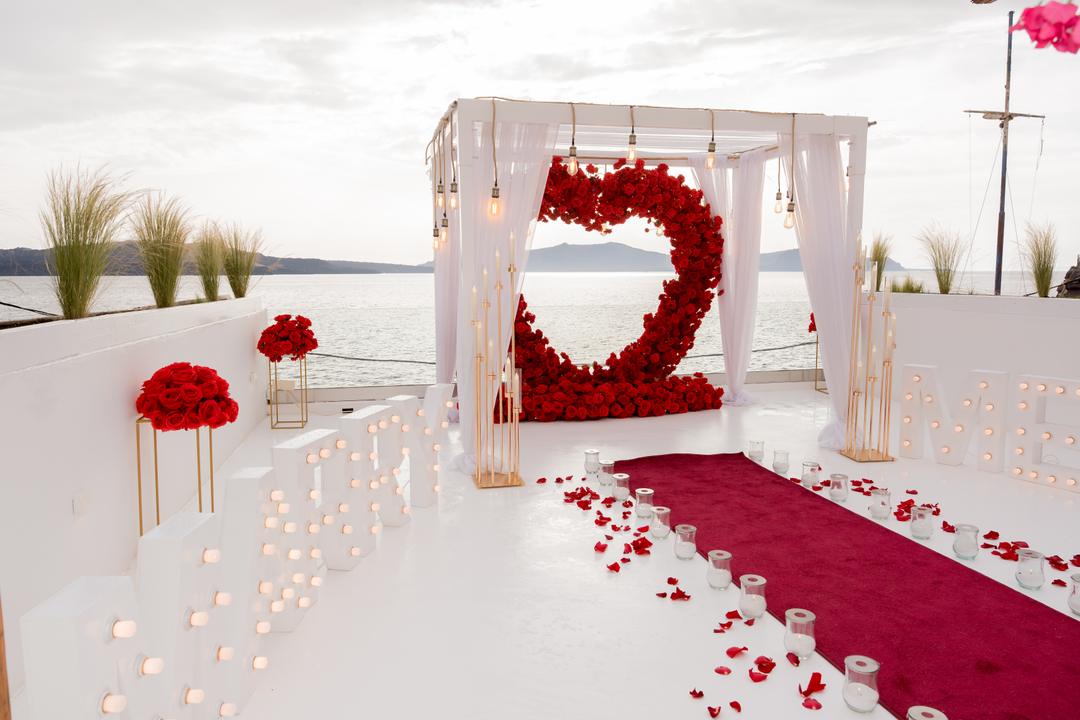 A red rose heart inside a white draped arch, a red carpet and long rows of candles leading to it across a white terrace above the sea