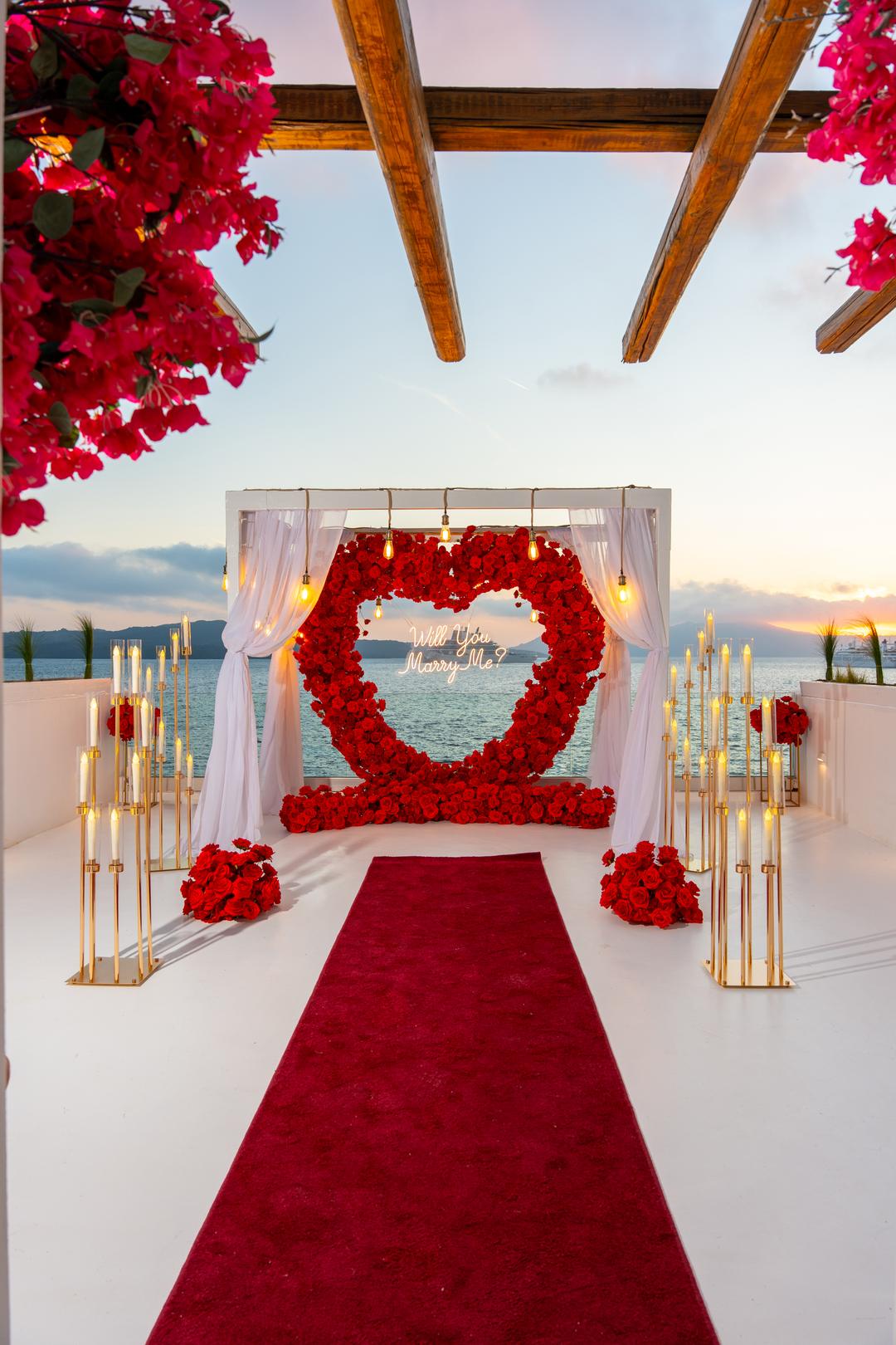 A red rose heart inside a white canopy at sunset, a red carpet and rows of gold candlesticks leading to it, bougainvillea overhead