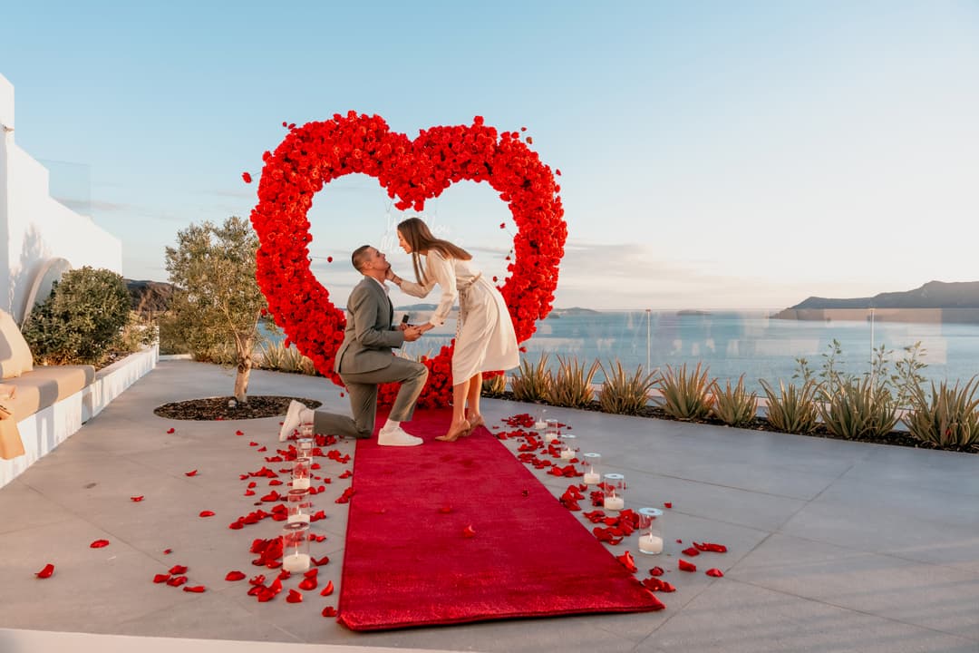 A man on one knee holding the ring box inside a tall red rose heart, candles and rose petals along a red carpet, the caldera and the sea behind them