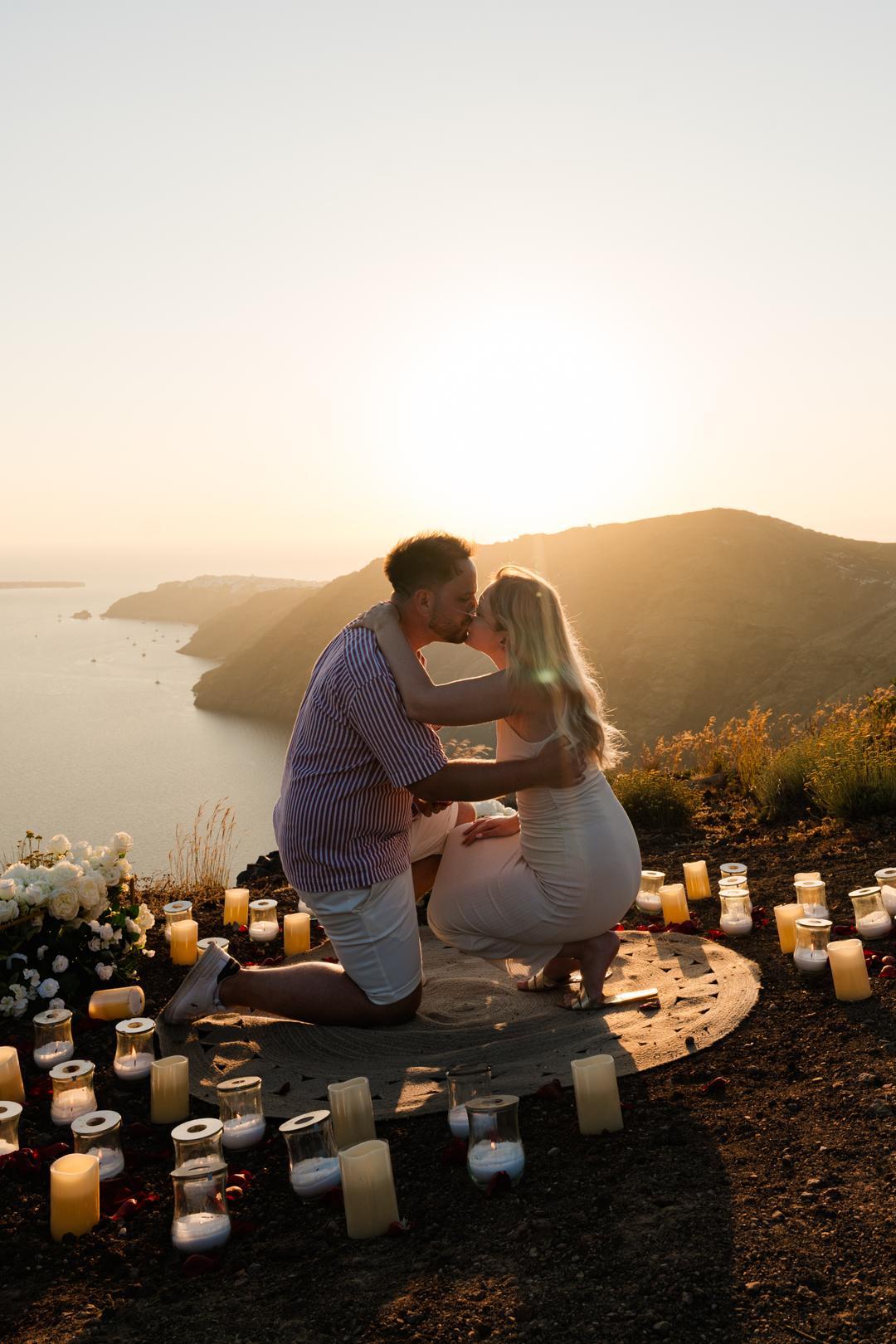A man on one knee on a rug ringed with candles at the cliff edge, kissing her hand as the sun goes down behind the caldera