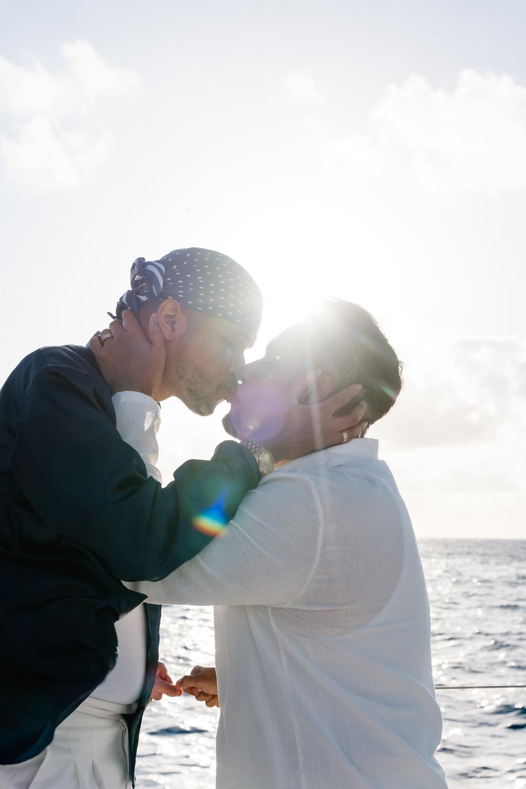 A couple leaning into a kiss on deck with the sun flaring behind them over open water