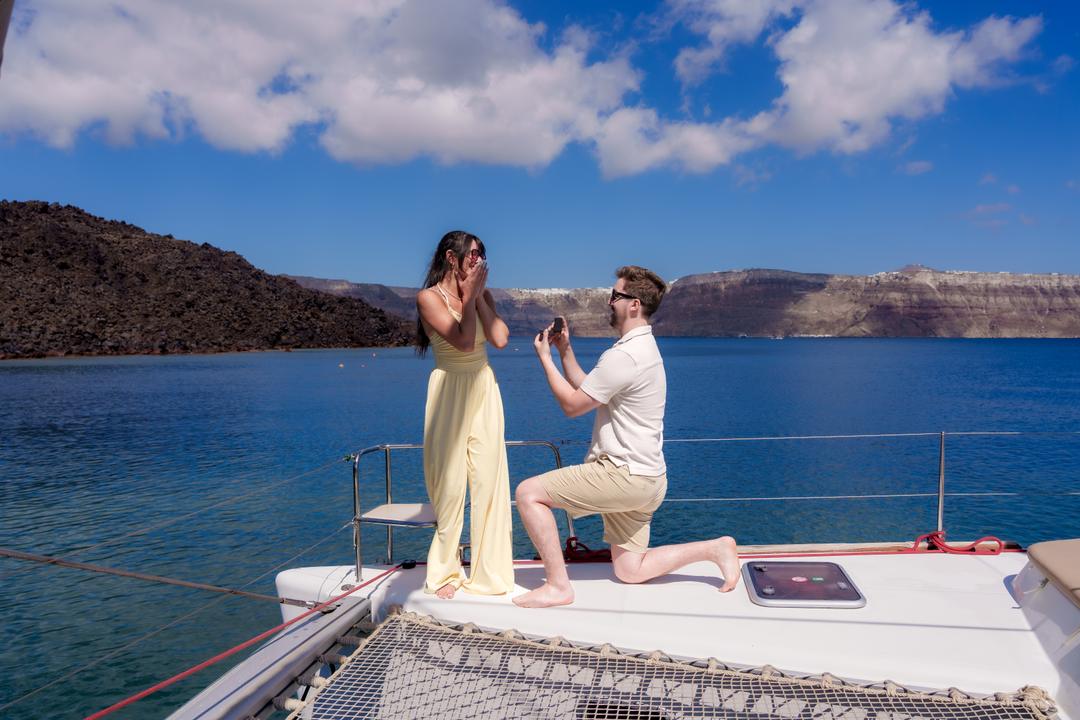 A man kneeling at the bow of a catamaran holding out a ring box, the volcano and the caldera wall behind on bright blue water