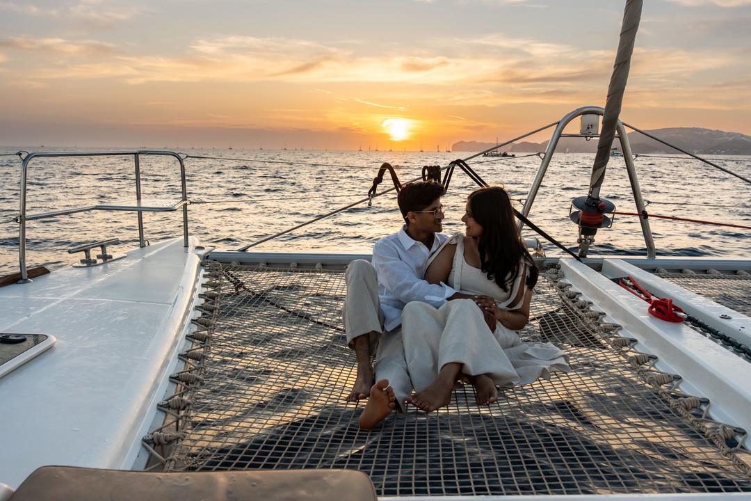 A couple sitting together on the netting between a catamaran’s hulls as the sun goes down over the water