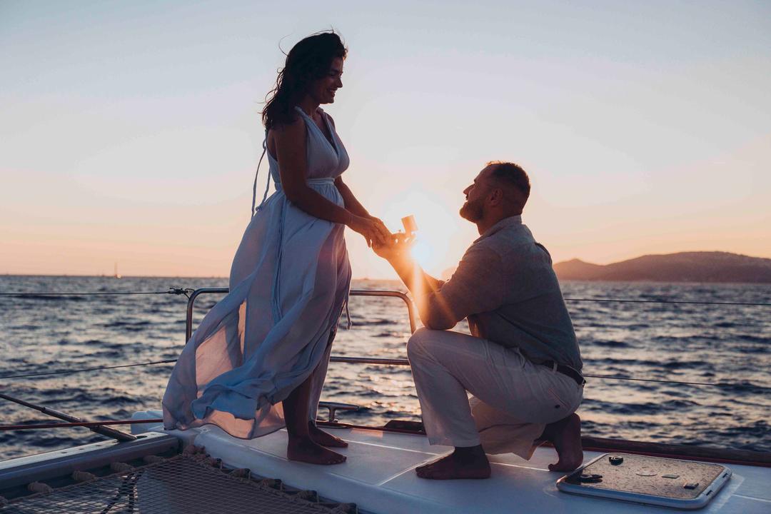 A man on one knee on the bow of a catamaran, the setting sun exactly between their joined hands and her dress lifting in the wind
