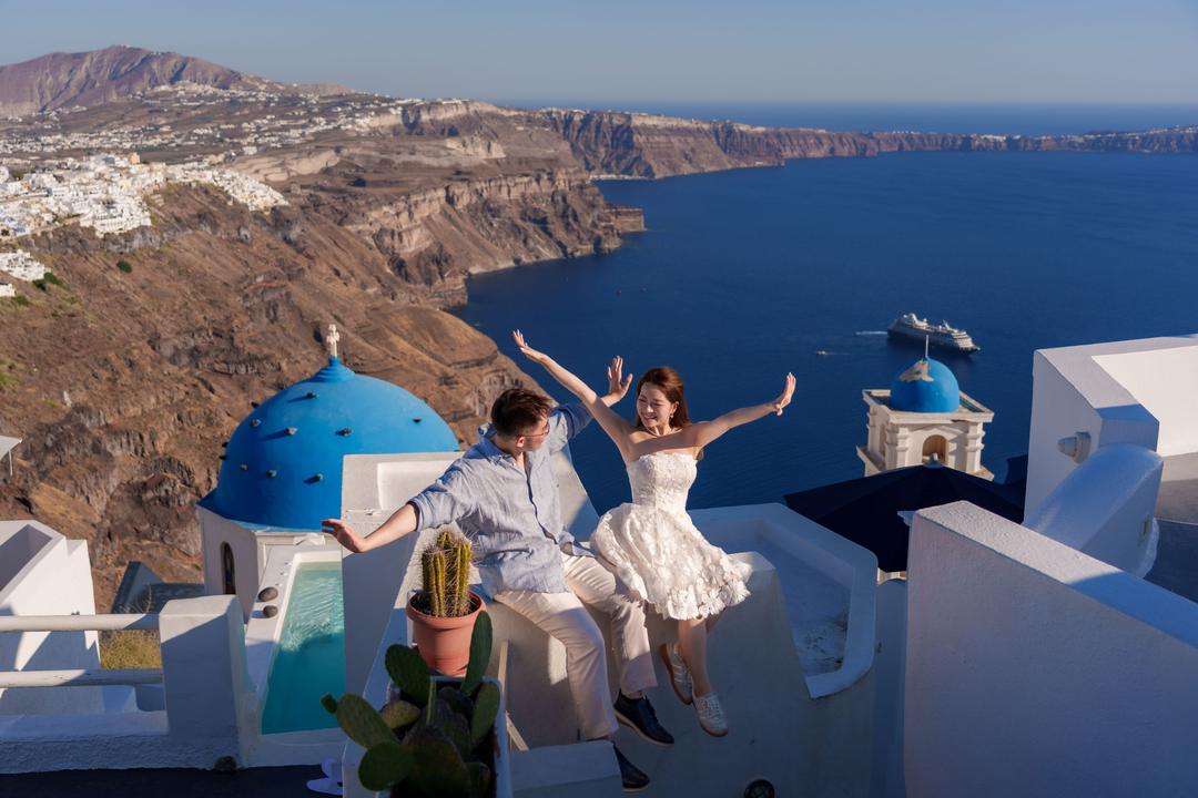 A couple with their arms thrown wide on a white terrace above two blue church domes, the caldera and a ship far below