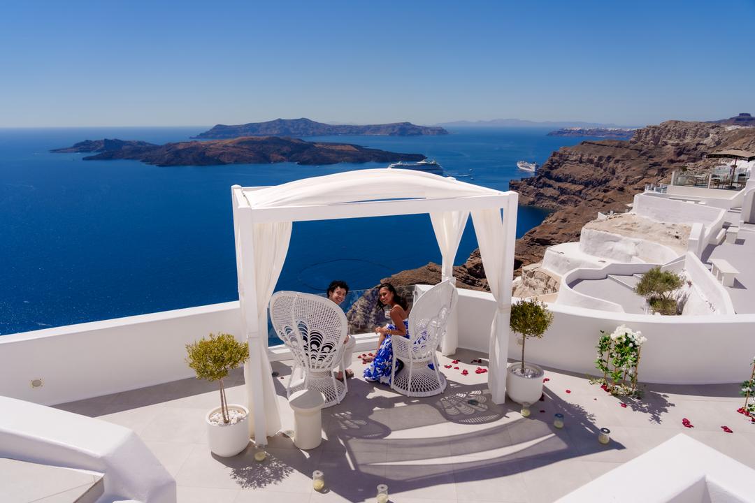 A white canopy with two chairs on a terrace, a couple seated under it, the caldera and the volcano islands filling the view