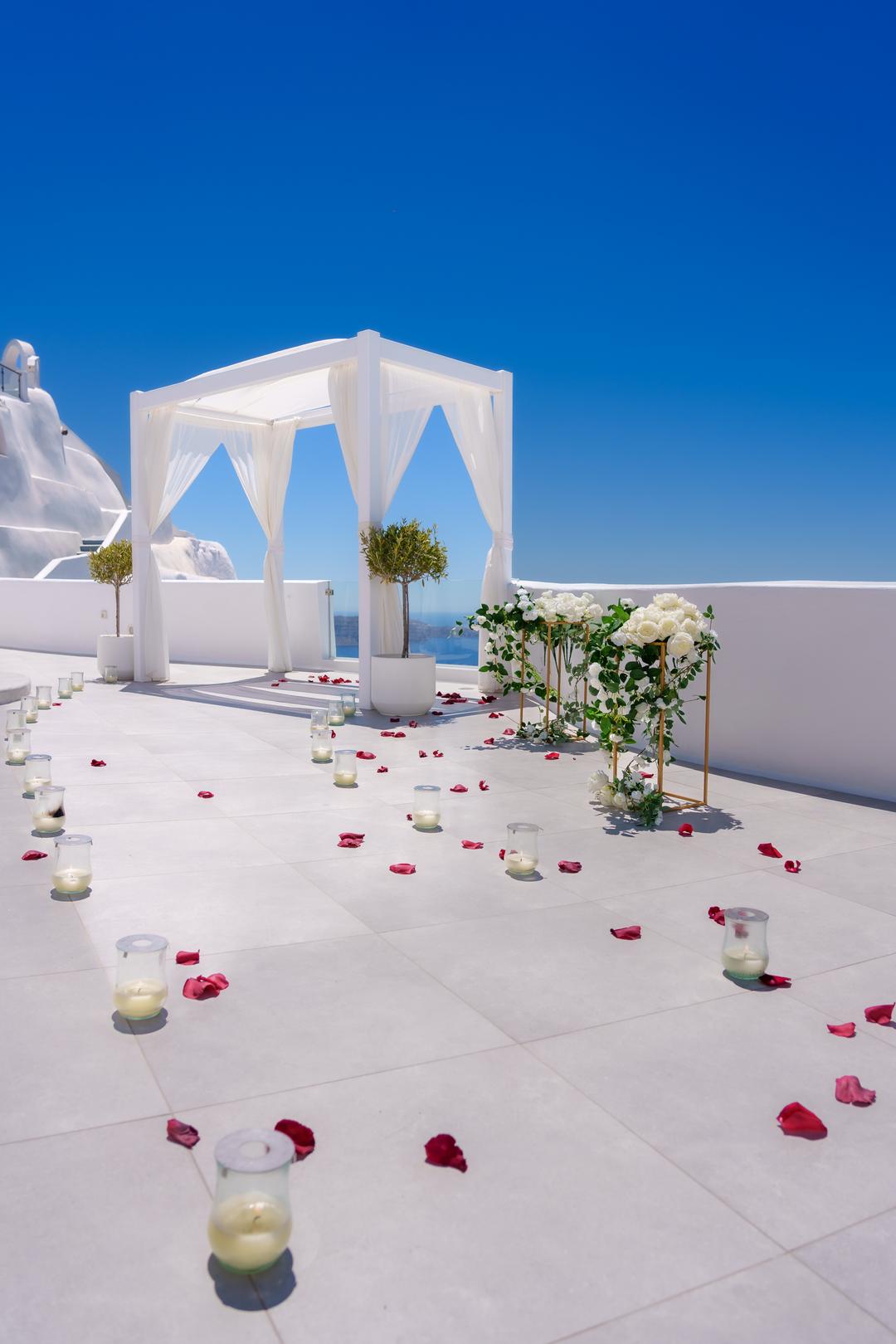 A white canopy on an empty terrace with a stand of white roses, glass candle holders and scattered rose petals under a deep blue sky