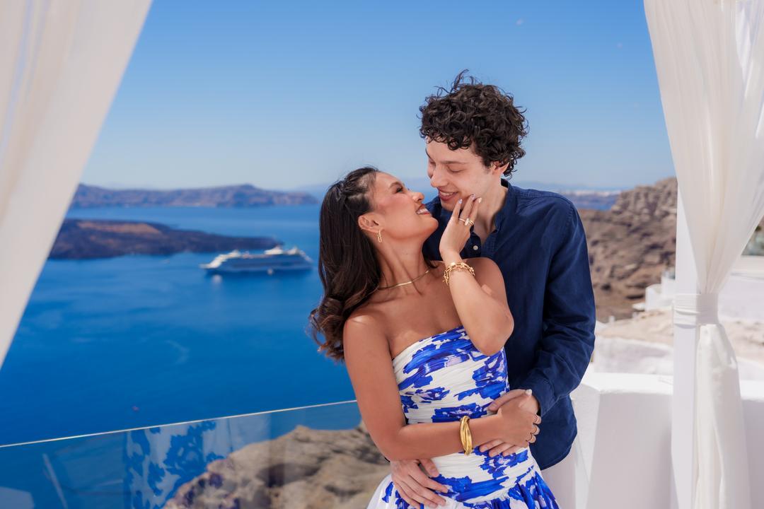A couple embracing between white drapes, the caldera, the volcano and a cruise ship behind them