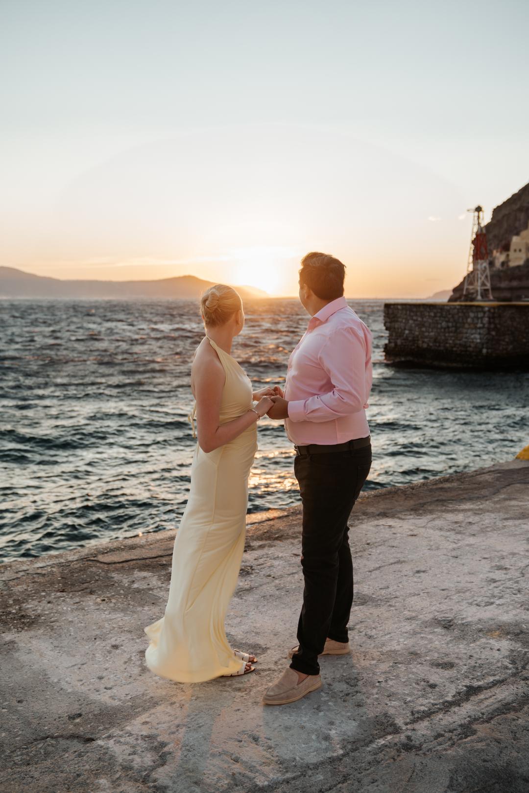 A couple standing on the rocks at the water’s edge with the sun low over the sea behind them