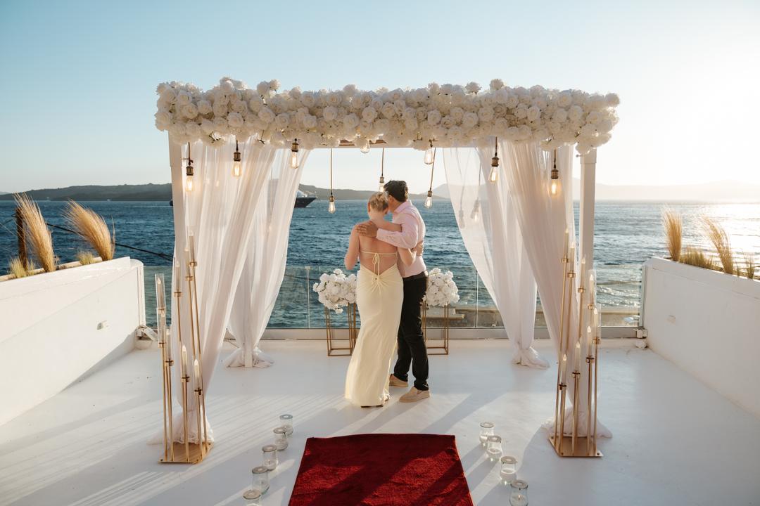 A couple embracing under a white floral arch set at the end of a jetty over the water, a red carpet and candles leading to it