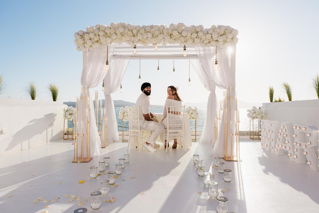 A couple under a white floral arch hung with filament bulbs, rose petals and candle glasses across the terrace floor