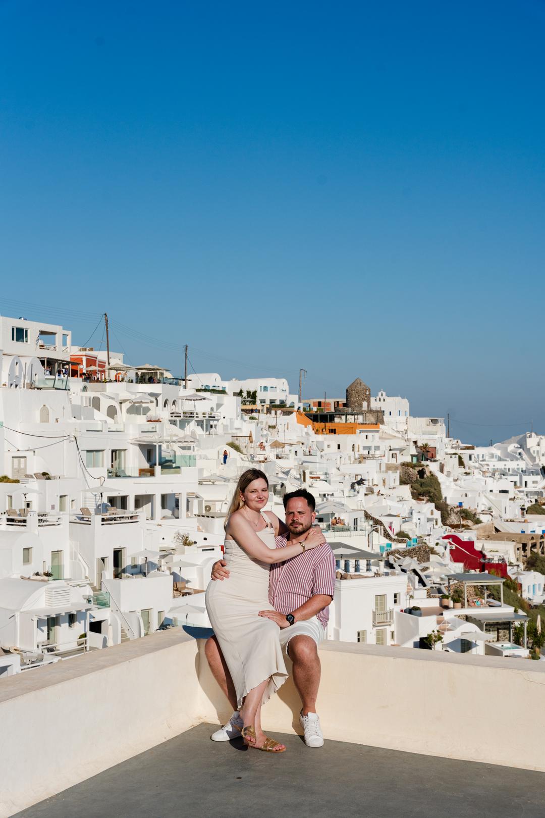 A couple on a rooftop terrace with a whitewashed village stacked down the cliff behind them