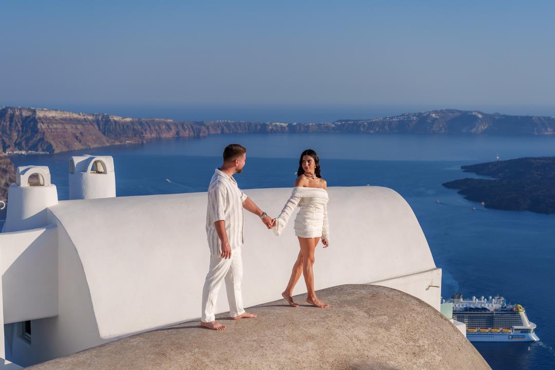 A couple walking barefoot hand in hand along a curved whitewashed rooftop above the caldera, a cruise ship on the water far below