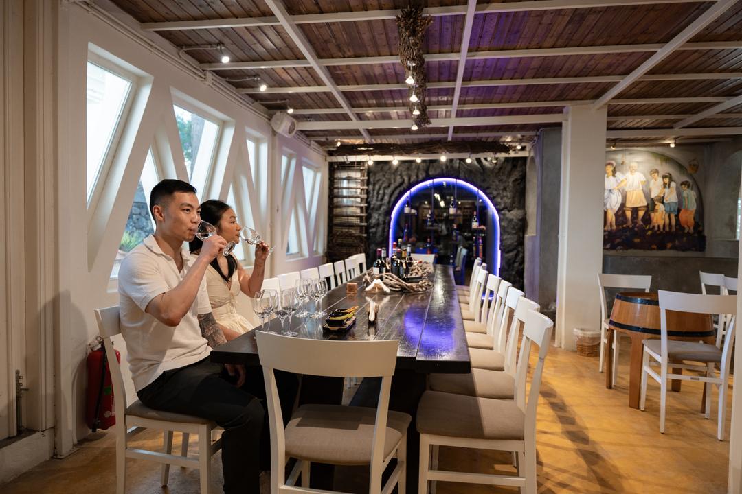 A couple tasting wine at the long table of a Santorini winery, tall windows down one wall and barrels at the far end