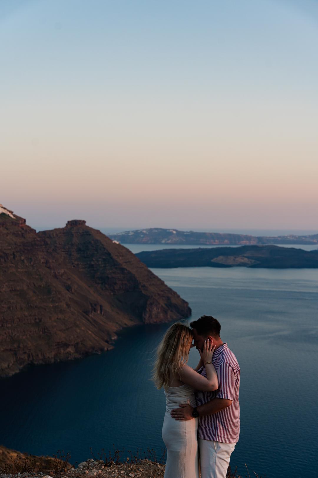 A couple embracing on the rim at dusk with Skaros rock and the still water of the caldera behind them