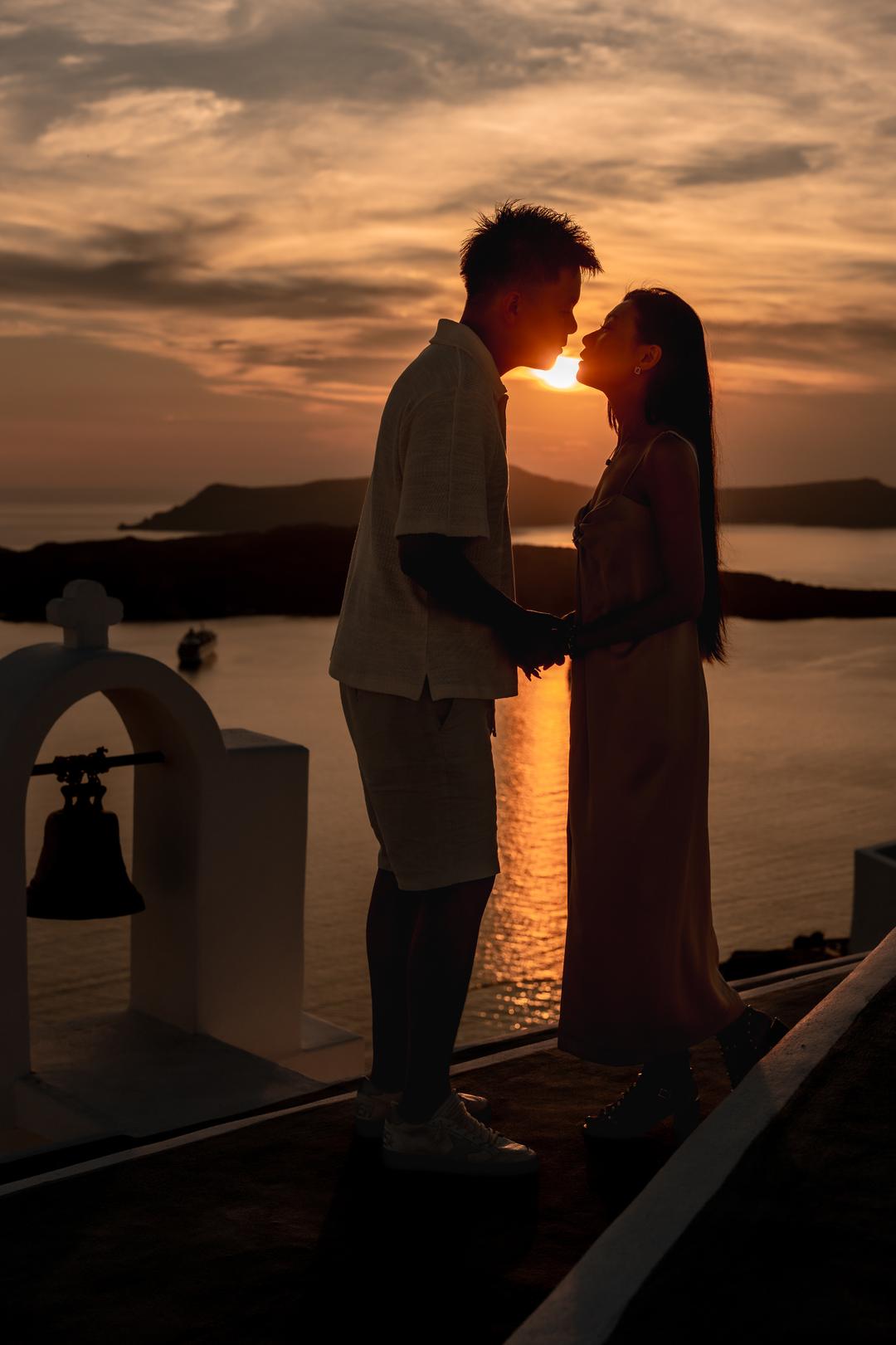 Two silhouettes beside a chapel bell as the sun lowers into the Aegean
