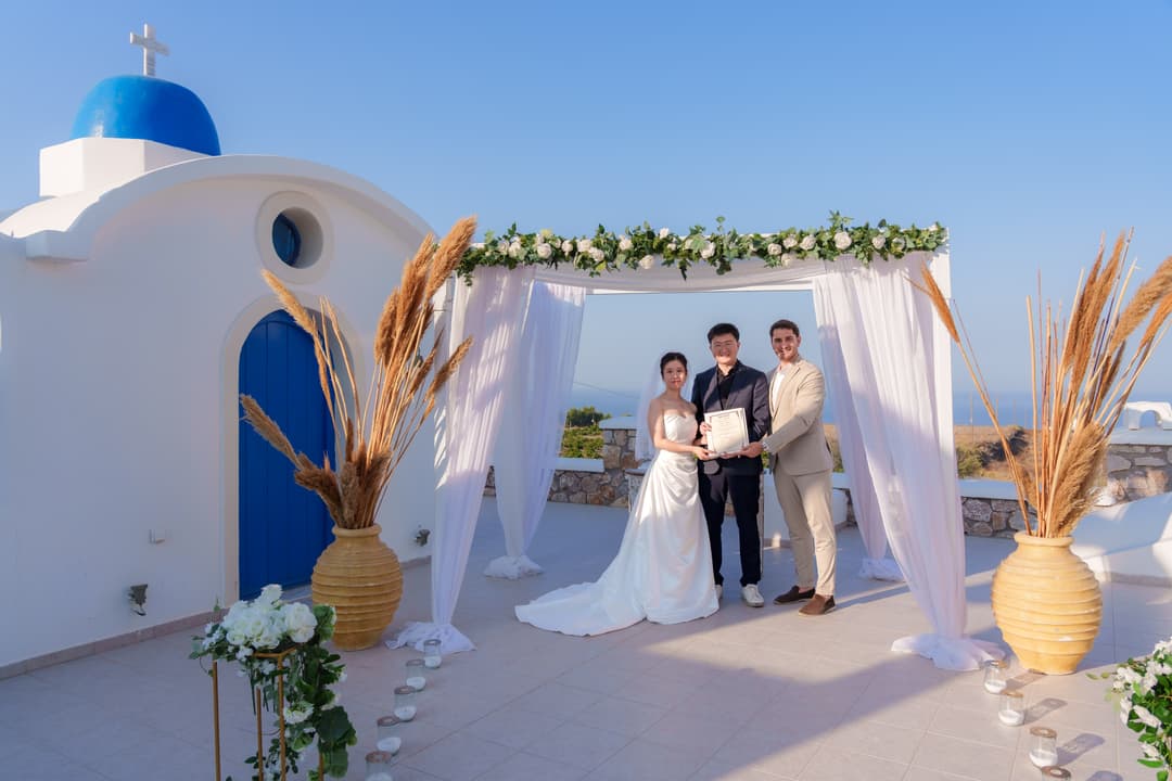 A couple and their celebrant holding the ceremony certificate under a draped canopy of white roses and pampas beside a blue-domed chapel