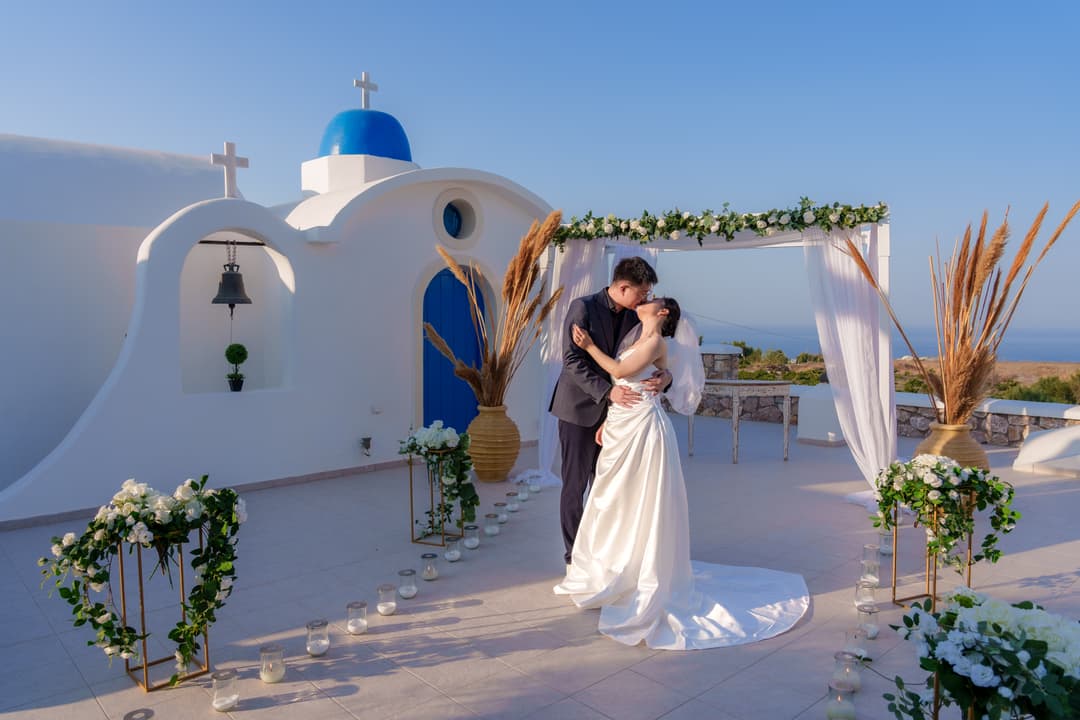 A bride and groom kissing beside their ceremony canopy, a white bell arch and blue-domed chapel behind them, candles along the aisle