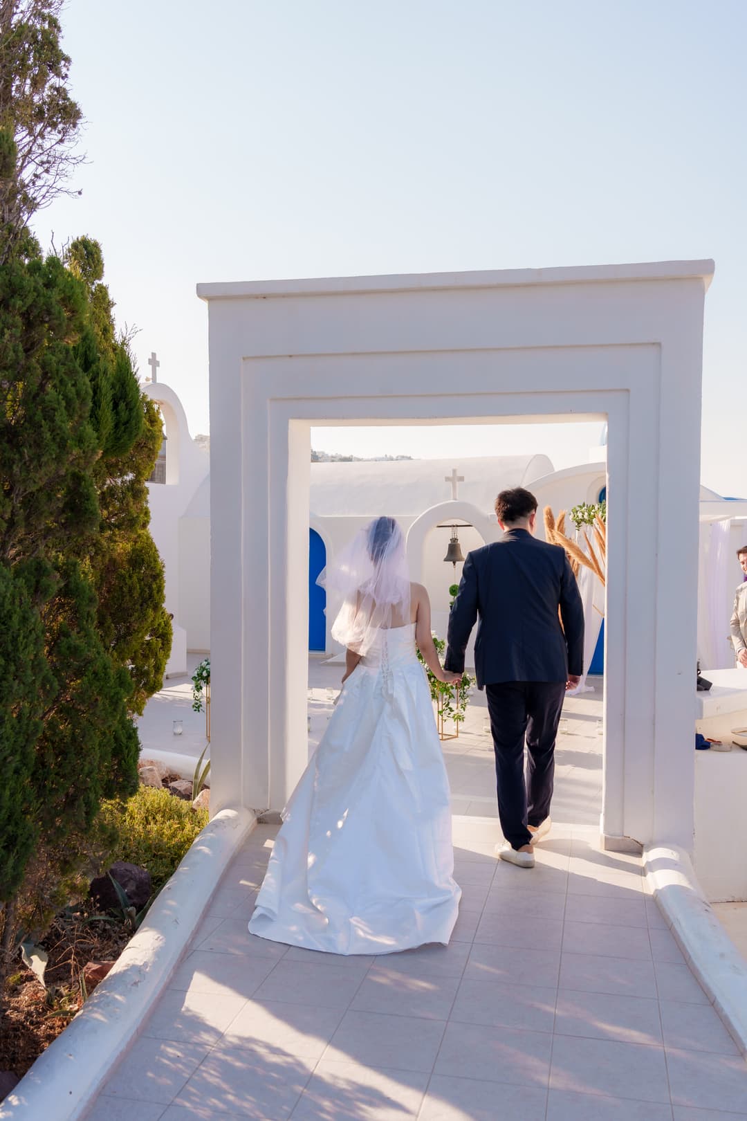 A bride and groom from behind, hand in hand, walking through a white gate toward a chapel courtyard dressed for their ceremony