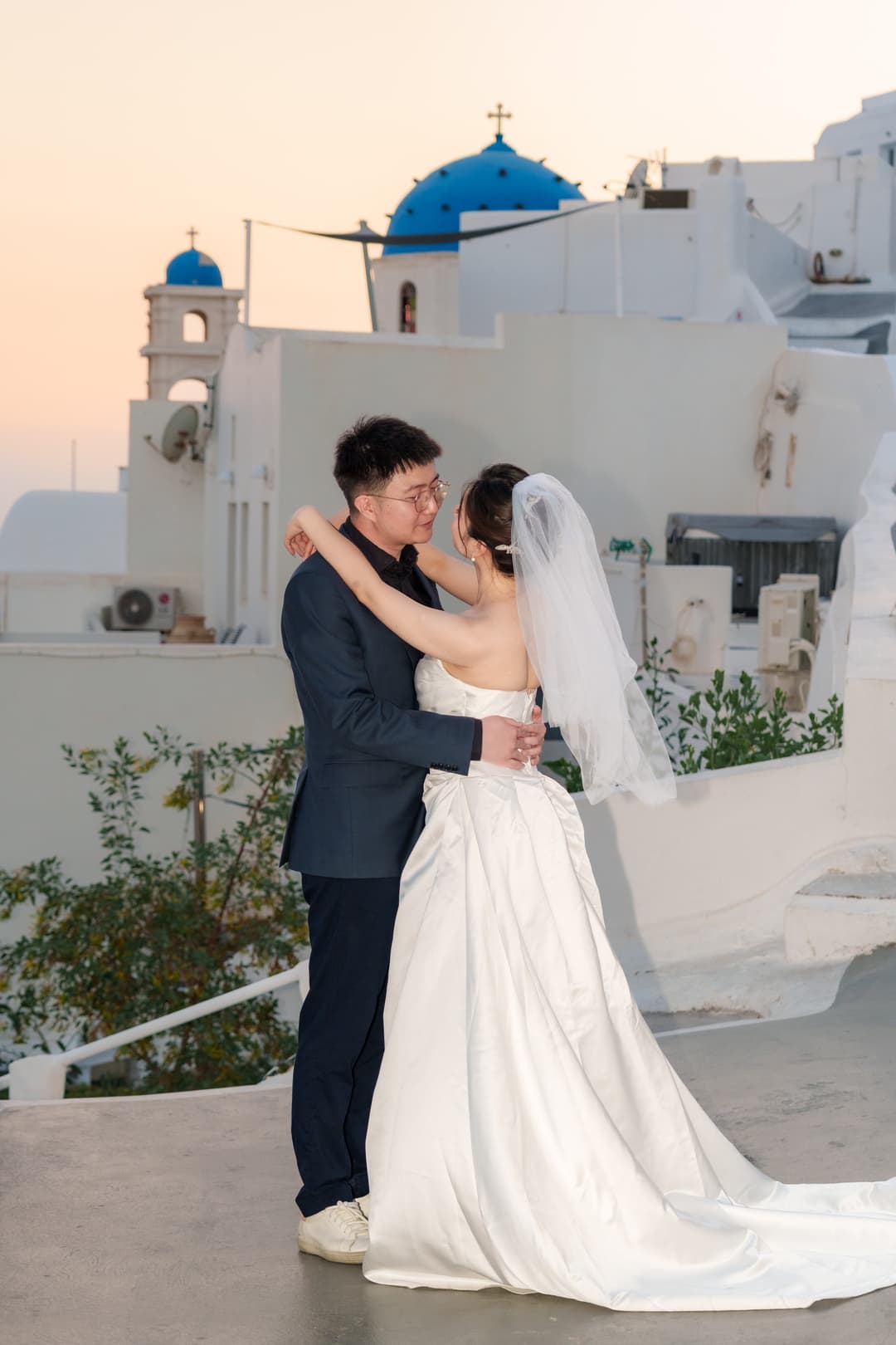 A couple holding each other at dusk, two blue-domed churches and the first pink of the afterglow behind them