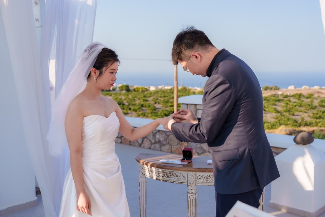 A groom placing the ring on his bride's hand at a small ceremony table, vineyard rows and the sea behind them